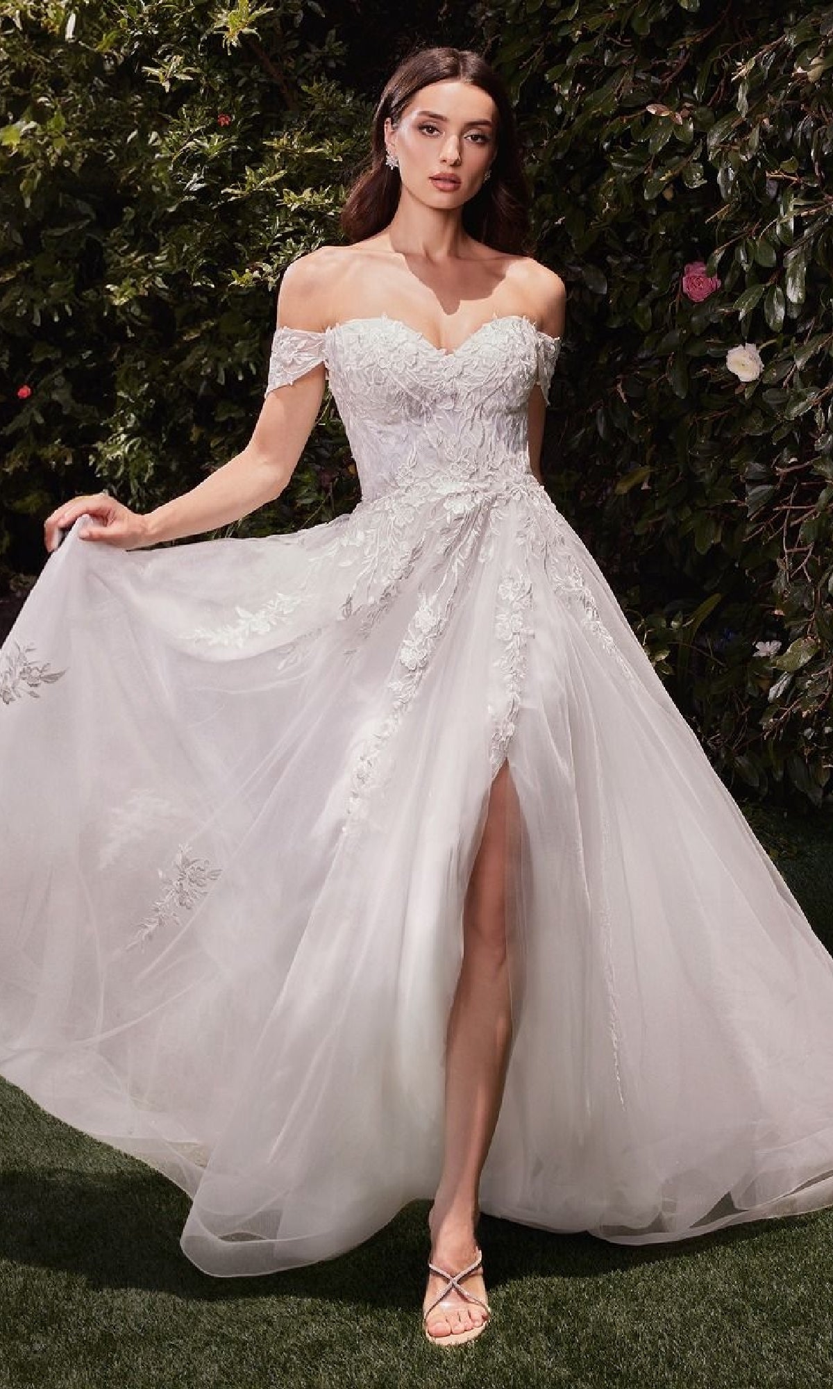 A woman wearing the Ladivine C159W off-white long wedding dress with lace and a slit stands on grass before lush greenery—an elegant, affordable gown perfect for your special day.